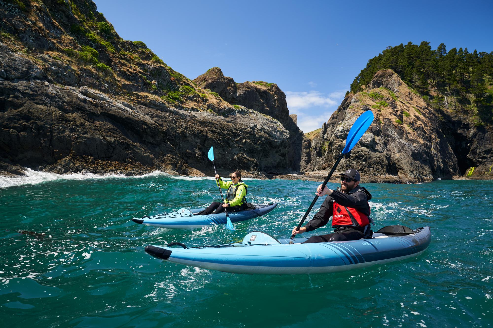 Two people kayaking in clear blue water surrounded by rocky cliffs and greenery.
