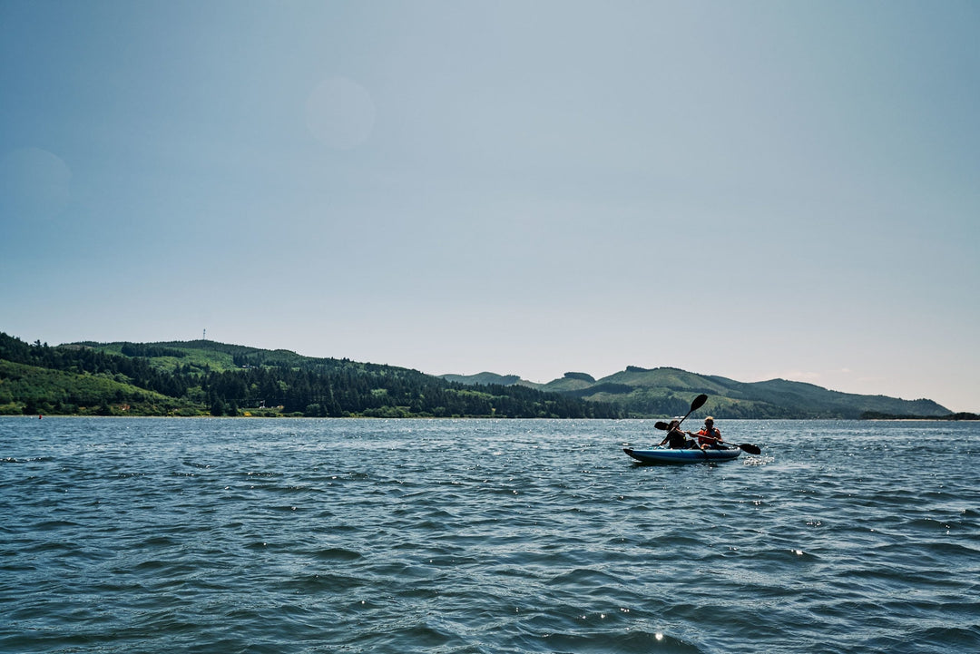 Two people kayaking in the water, with hills and trees in the background under a clear sky.