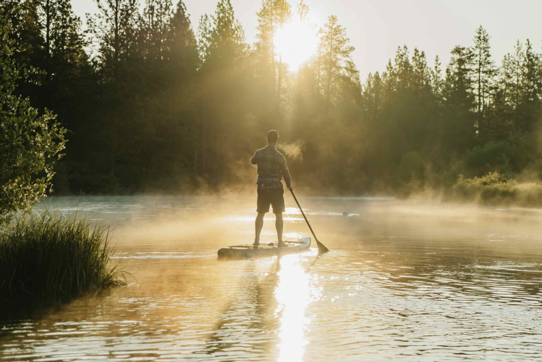 Person paddling on a misty lake at sunrise with trees and sun rays in the background.