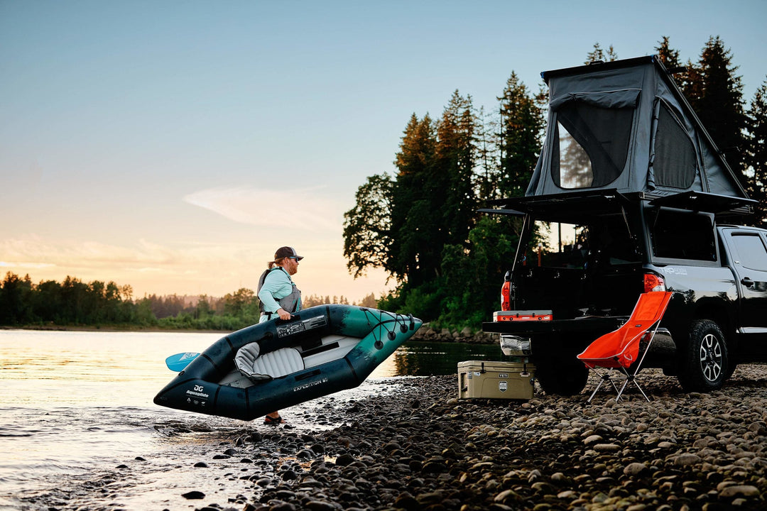 Man carrying a kayak near a truck with a rooftop tent and camping gear by the water.