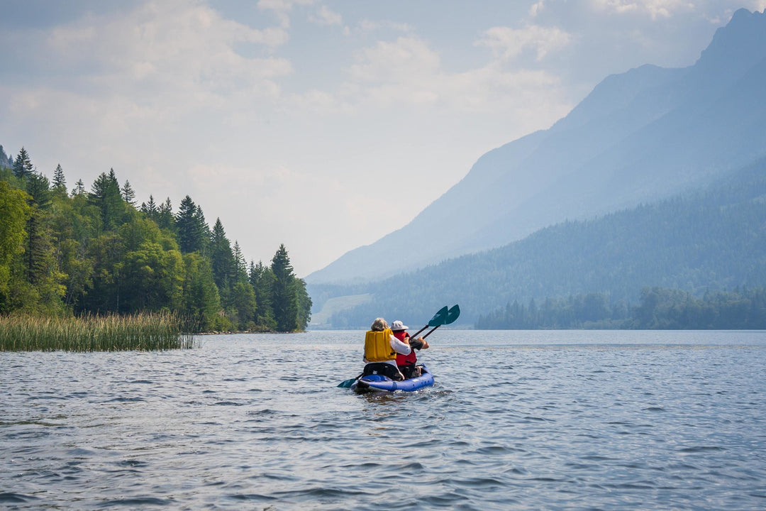 Two people paddling a kayak on a lake surrounded by trees and mountains.