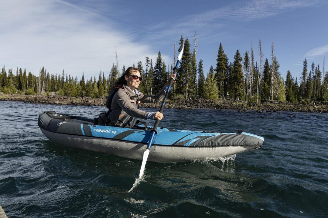 Person smiling while kayaking on a calm lake with trees and blue skies in the background.