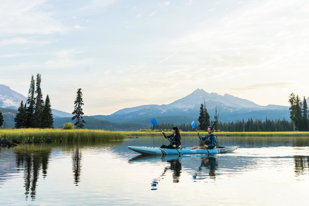Two people kayaking on a serene lake with distant mountains and evergreen trees.