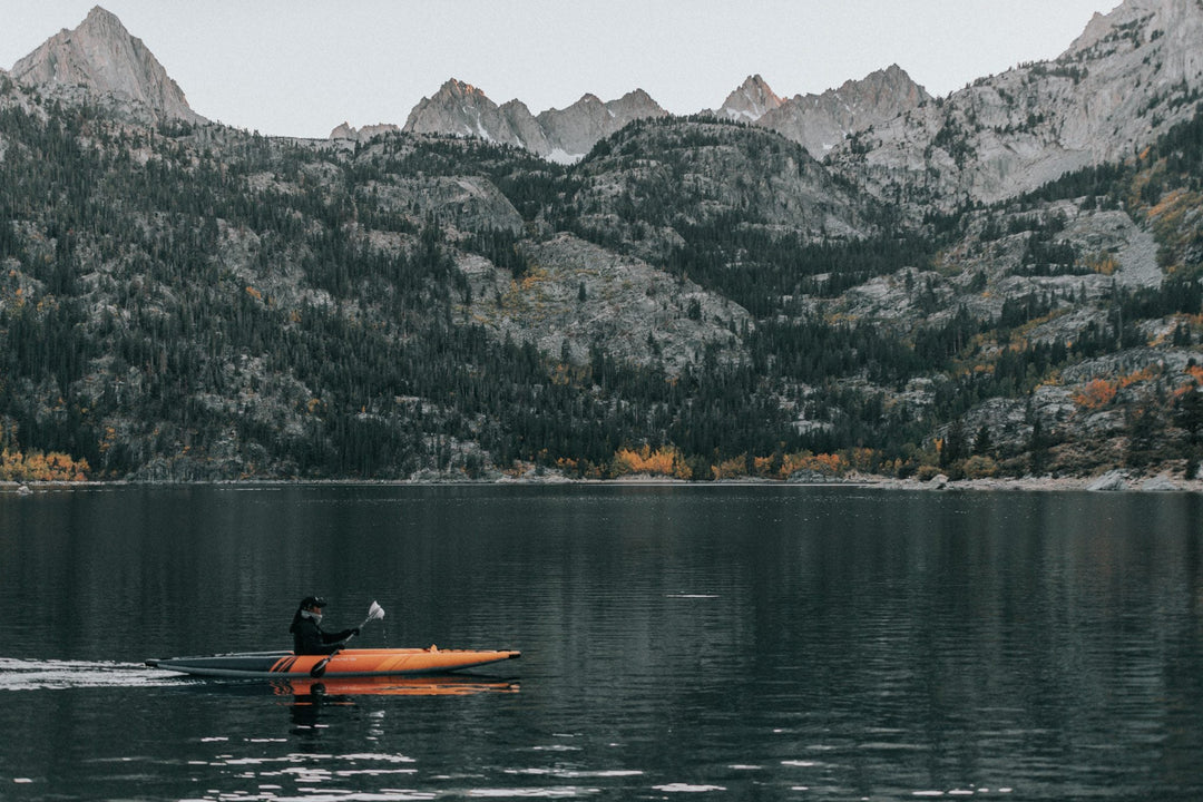 Person in an orange kayak on a calm lake with mountains in the background.