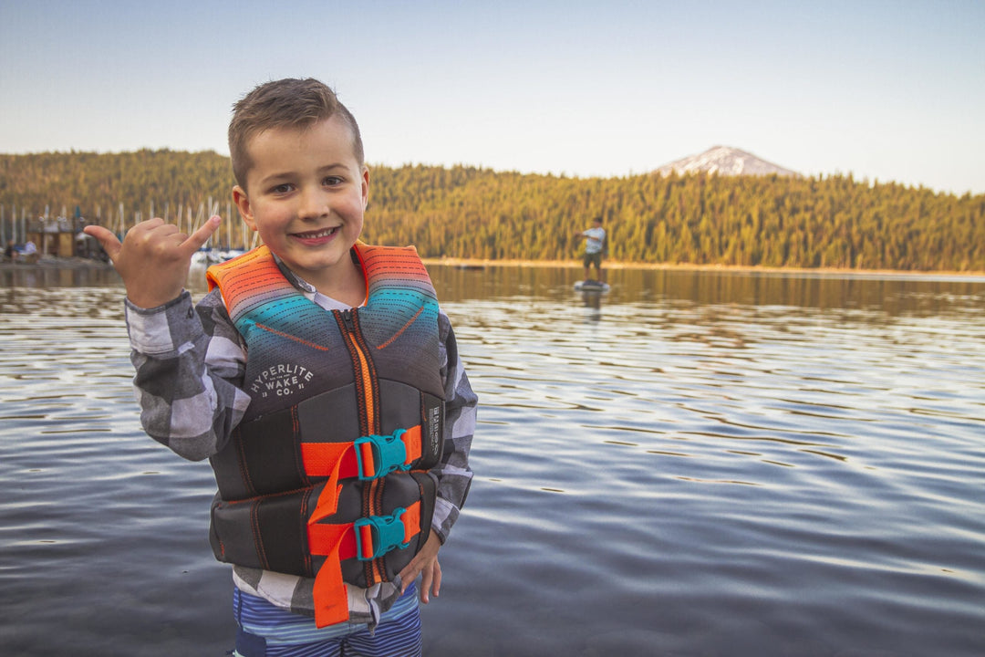 Boy in life jacket giving a thumbs-up with a mountain backdrop and paddleboarder behind.