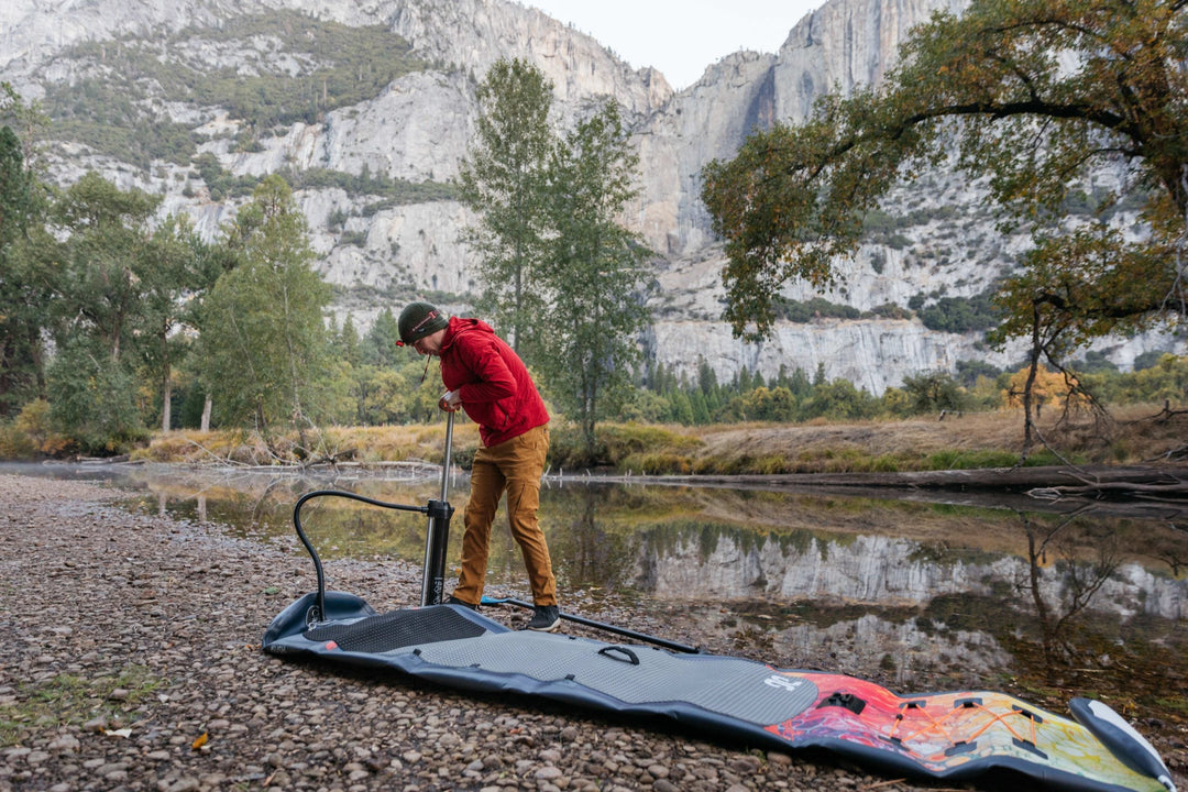 Person inflating a paddleboard by a river with rocky terrain and mountain background.