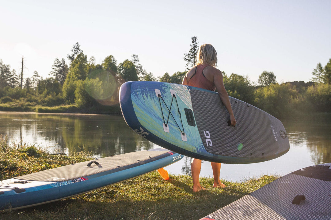 Woman carrying a paddleboard near a calm lake, with trees and clear skies in the background.