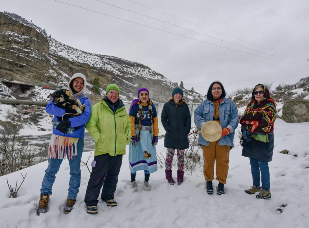 Group of six people and a dog standing in the snow with mountains in the background.