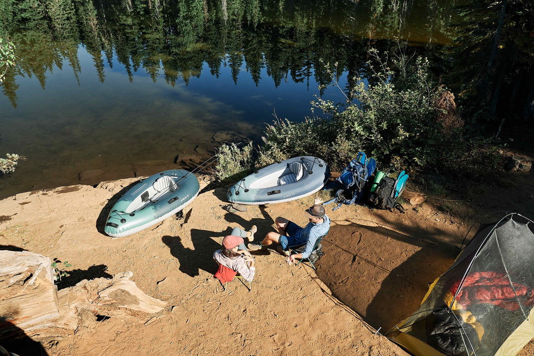 Two inflatable kayaks on a riverbank next to backpacks and a tent in a forested area.