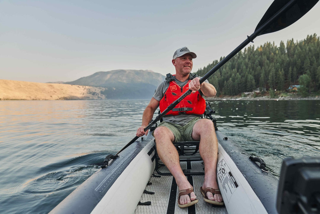 Man paddling a kayak on a calm lake with mountains in the background.