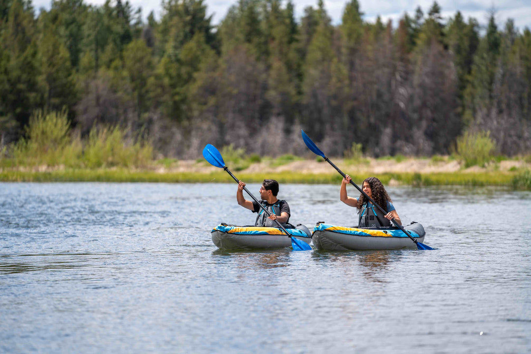 Two people kayaking on a calm river with green trees and a blue sky in the background.