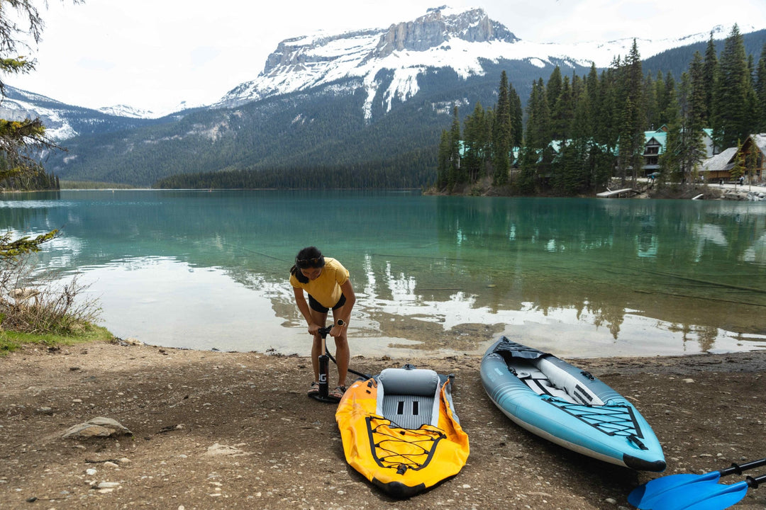 Person pumping air into a yellow inflatable kayak by a calm, clear lake with mountains.