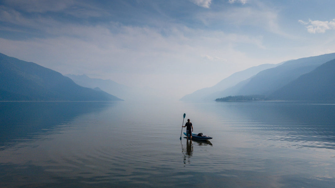 Man stands on a paddleboard on a still lake with mountains in the background.