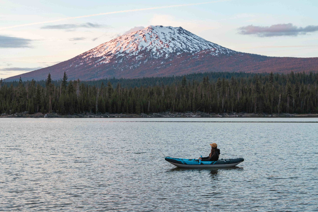 Person kayaking in an inflatable kayak on a calm lake at sunset with a snow covered mountain in the background.