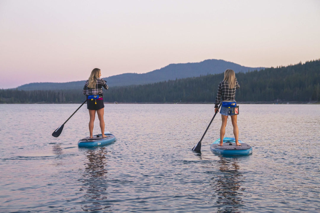Two women paddleboarding on a calm lake with mountains in the background at dusk.