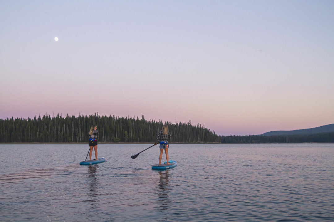 Two people paddleboarding on a lake at sunset with distant trees and soft pink hues in the sky.