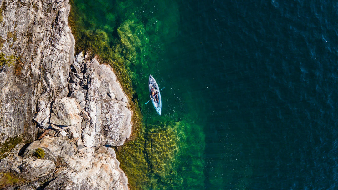 Aerial view of a person kayaking near a rocky shoreline, with clear green and dark blue water.