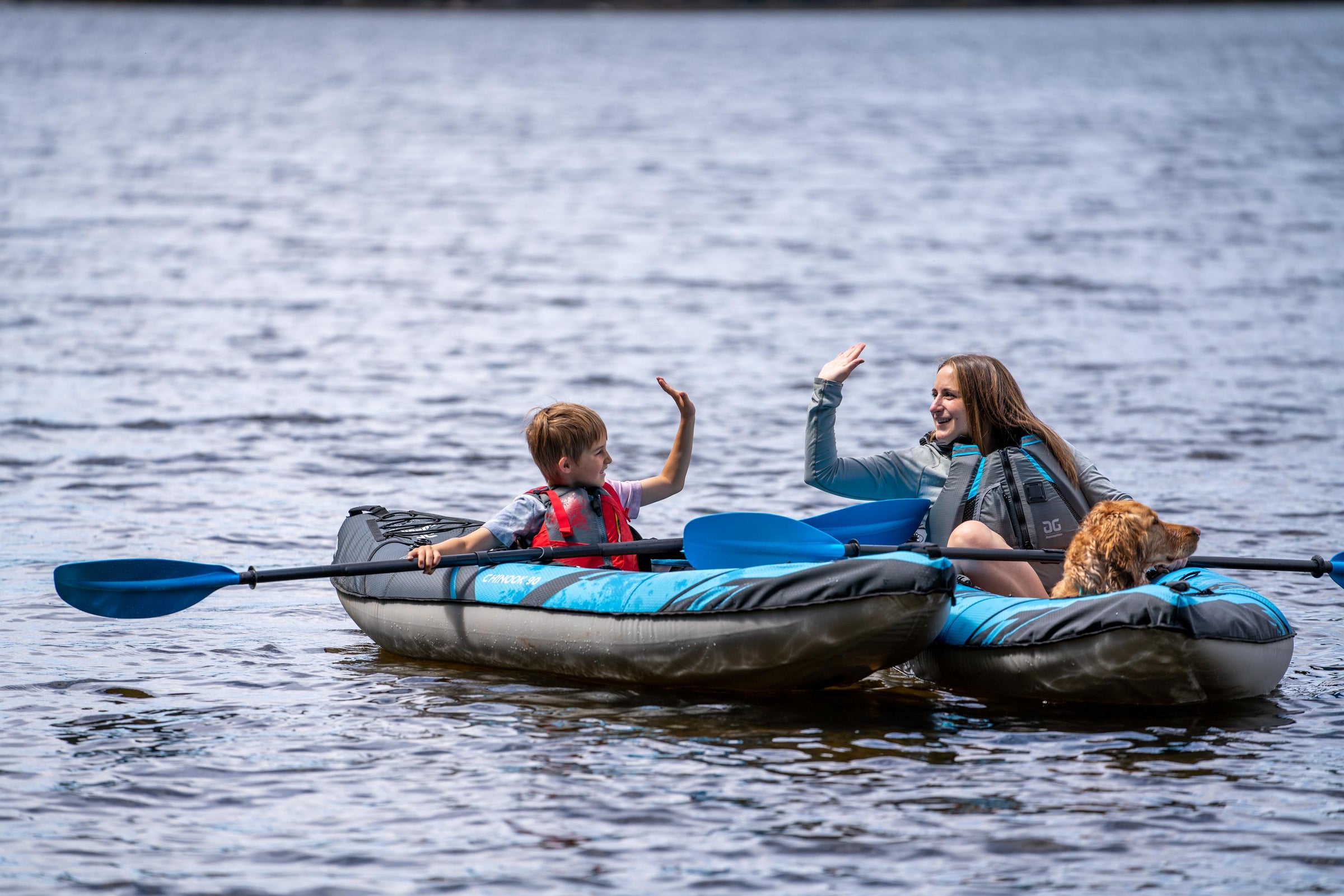A woman and child in kayaks high-five on a lake, with a dog sitting in one kayak.