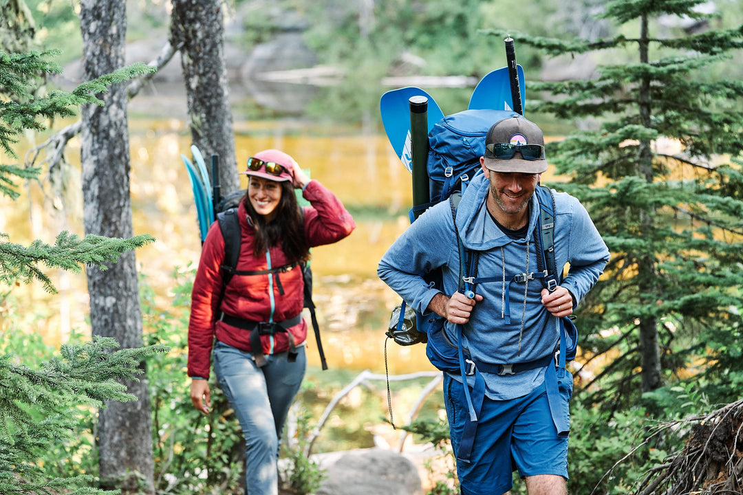 Two people hiking in the forest near a body of water, carrying backpacks and smiling.
