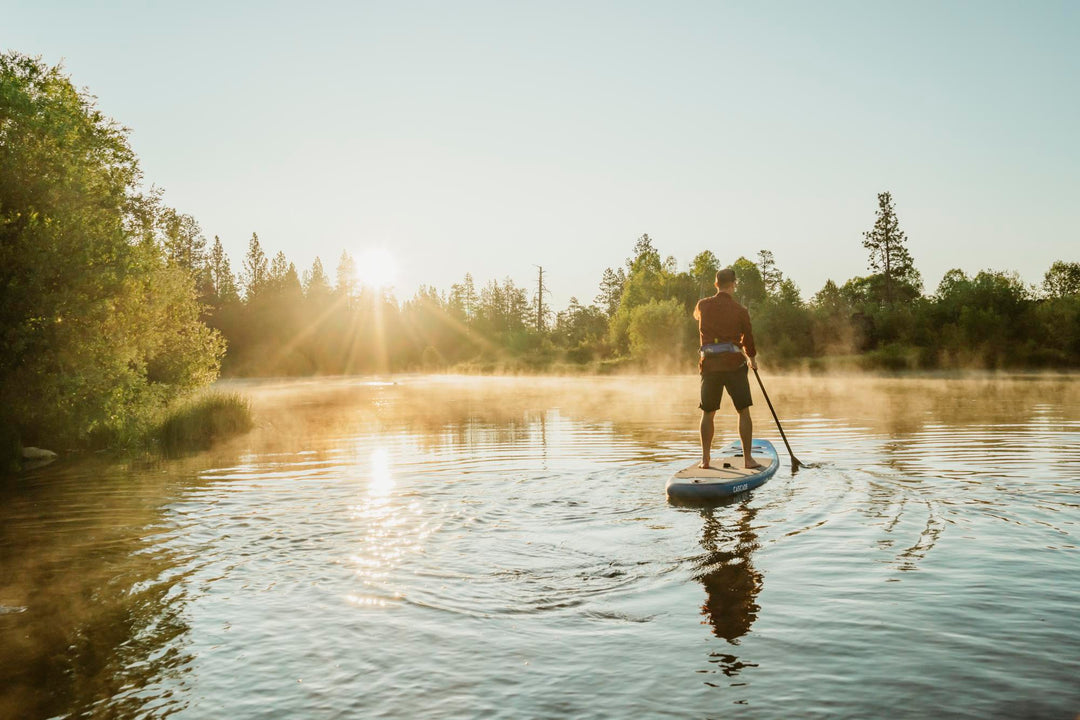 Person paddleboarding on a misty lake at sunrise, surrounded by trees.