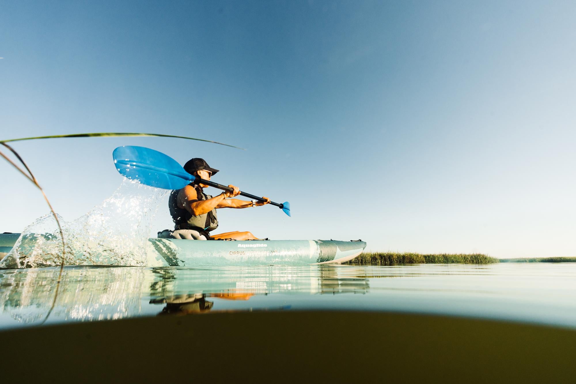 Person kayaking on calm water, creating a splash with a blue paddle under a clear blue sky.
