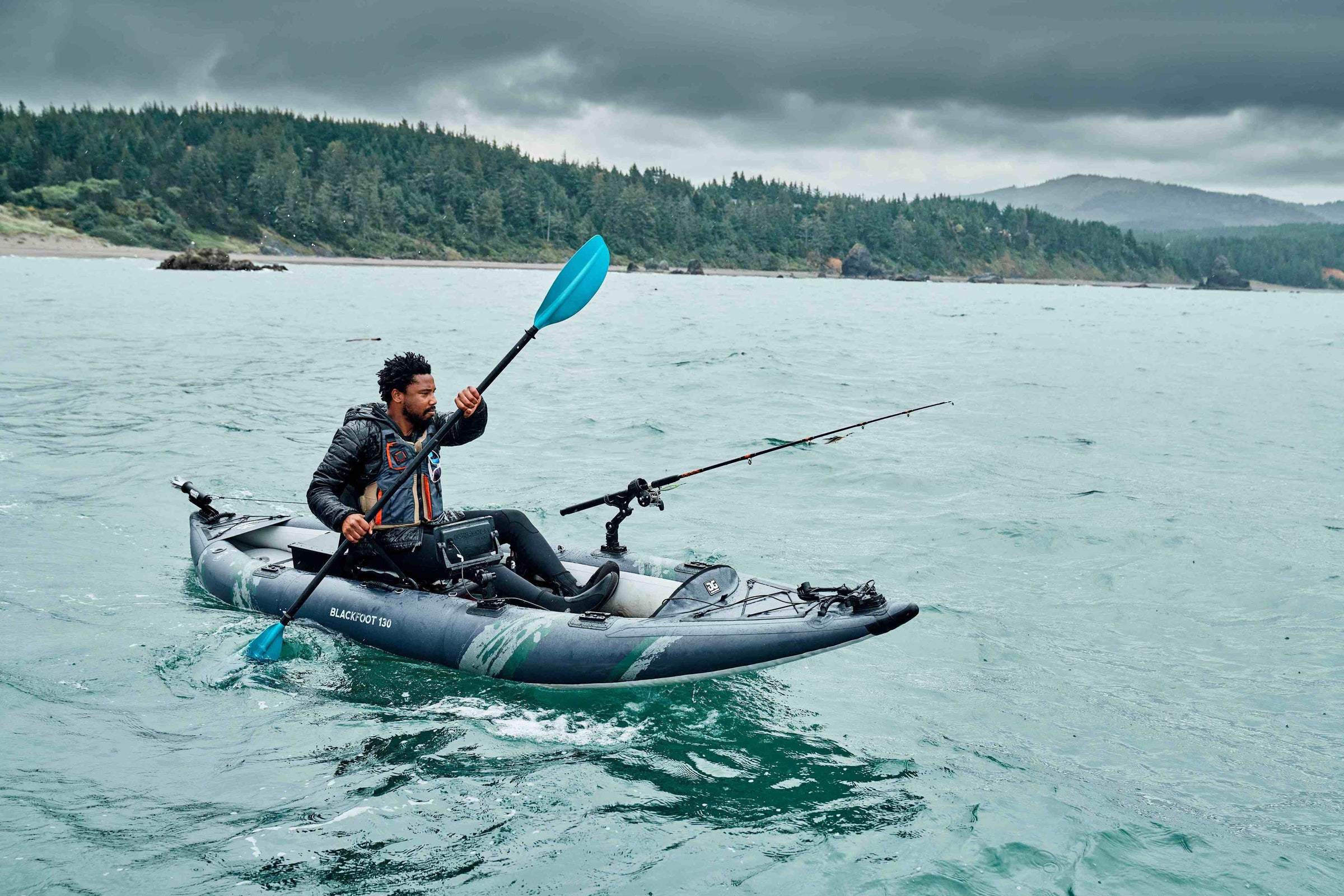 Person kayaking and fishing on a calm sea under a cloudy sky, near a forested coastline.