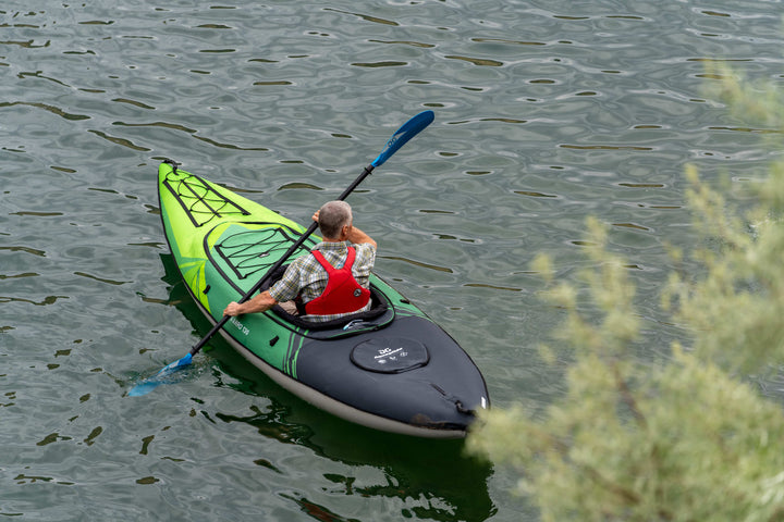 A person kayaking in a Navarro 130 on calm water, with greenery in the foreground.