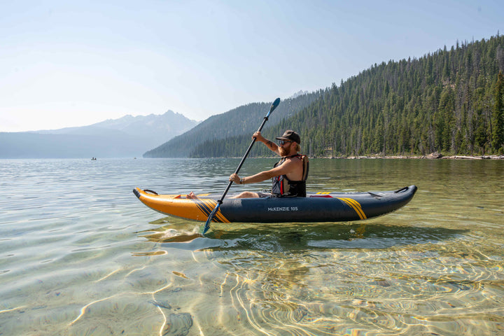 Paddling a McKenzie 105 kayak on a tranquil lake with forested mountains in the backdrop.