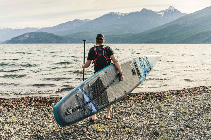 Roam 126 Paddleboard user stands on rocky lakeshore with mountains in view.