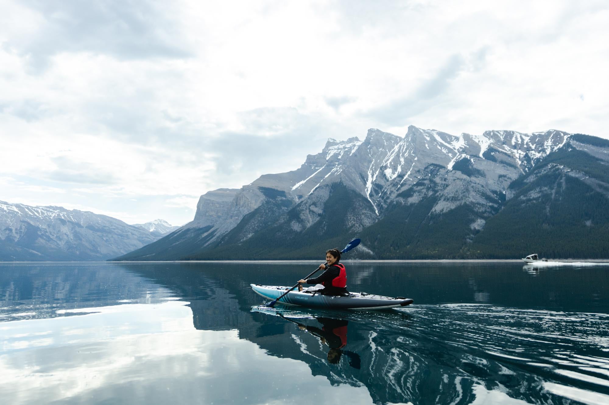 Person kayaking on a serene lake with snow-capped mountains in the background.