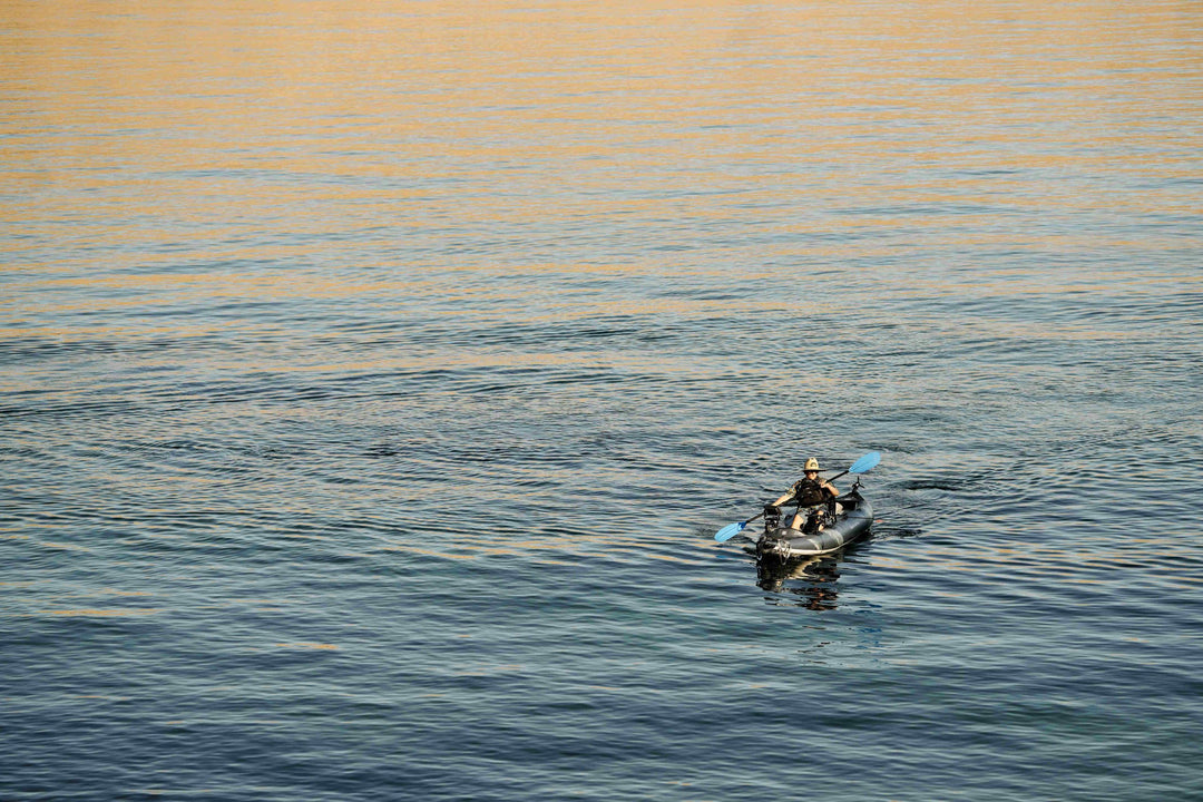 Person kayaking on calm water in a Blackfoot Angler 130 under a clear sky.