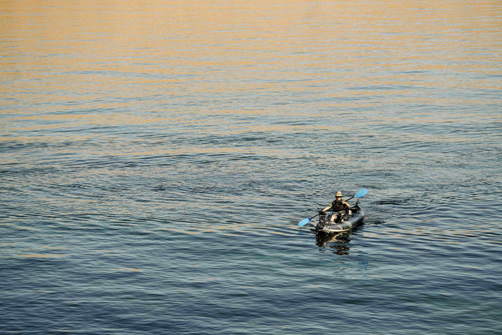 Person kayaking on calm water in a Blackfoot Angler 130 under a clear sky.