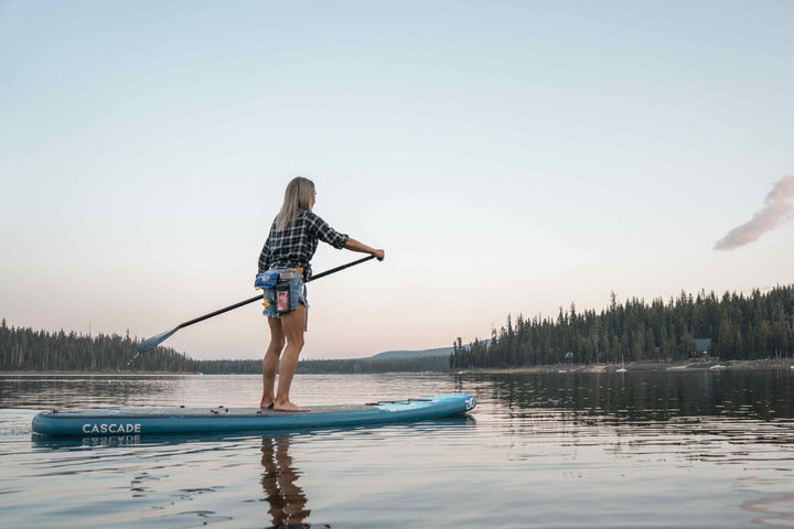 A paddleboarder showcases the sleek Cascade 10 Paddleboard Package on a calm lake at sunset.