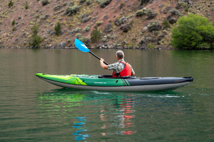 Man kayaking in a Navarro 130 on a calm lake by the rocky shore and greenery.