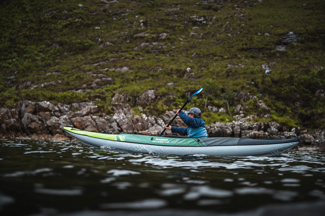 Person enjoying a calm lake paddle in the Navarro 145 inflatable kayak near a rocky shore.