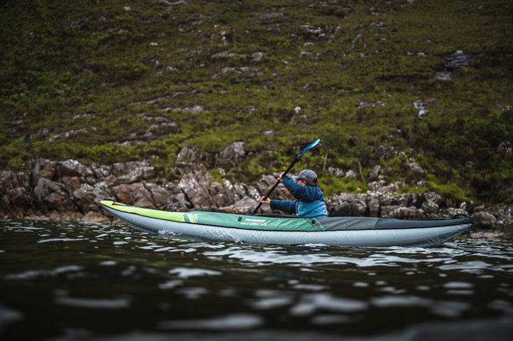 Person enjoying a calm lake paddle in the Navarro 145 inflatable kayak near a rocky shore.