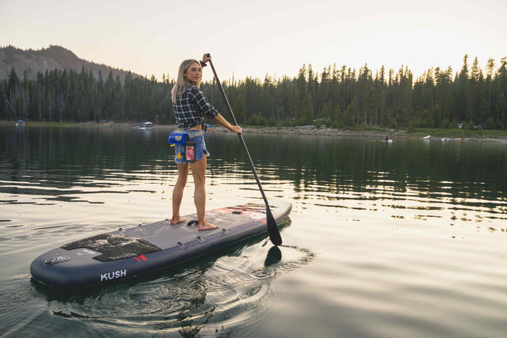 Woman paddles the Kush 11 on a calm lake at sunset, surrounded by trees and hills.