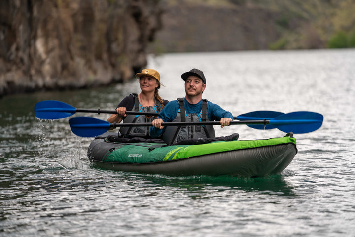 Two people paddle a Navarro 145 kayak on a lake with rocky cliffs around.
