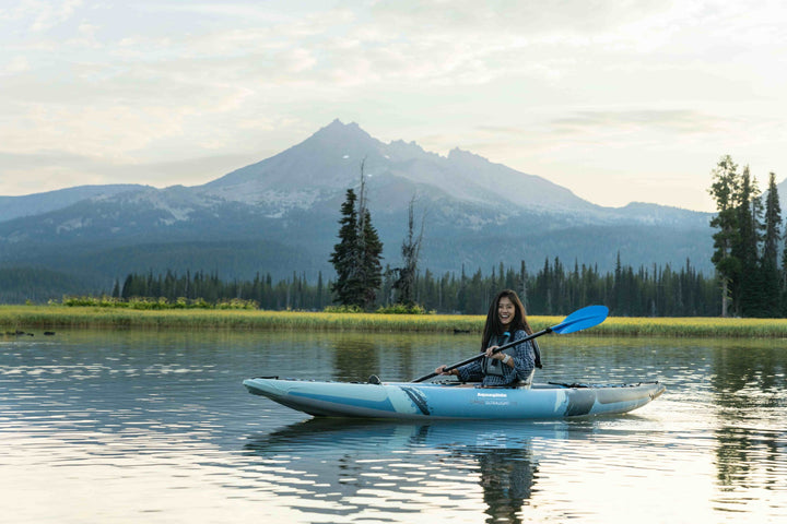 Person kayaking on a calm lake in a Cirrus Ultralight 110 among mountains and trees.