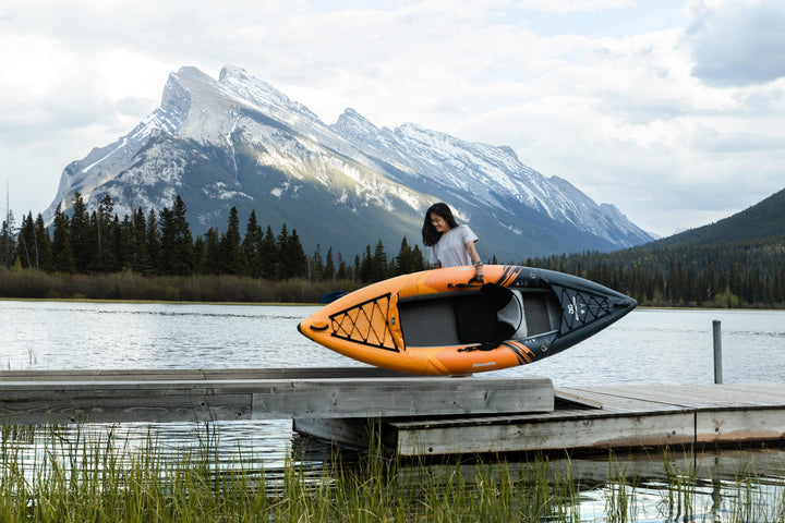 Individual carrying the Deschutes 110 kayak on a dock, mountains and trees beyond.