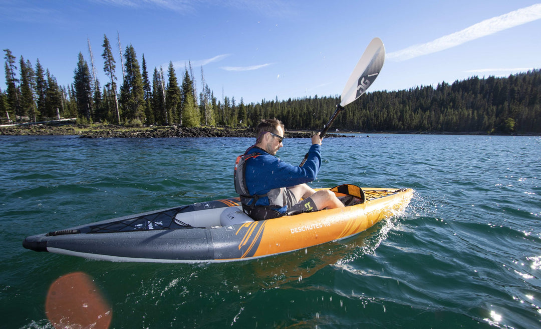 A person paddles the Deschutes 110 kayak on a serene lake under a blue sky.