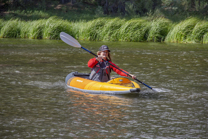 Individual using the Deschutes 130 Package kayak on a river amidst lush greenery.