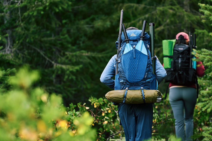 Two hikers with Backwoods Purist 65 backpacks trek a lush forest trail by an ultralight kayak.