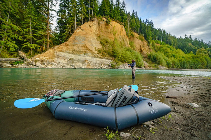 Backwoods Expedition 85 kayak on riverbank, person fly-fishing, surrounded by trees and cliffs.